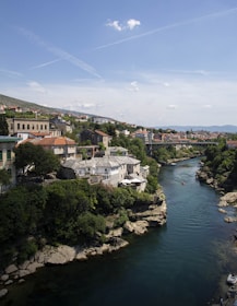 A picturesque view of a river flowing through a town with buildings featuring red-tiled roofs. Lush greenery flanks the water, while a bridge spans across the river in the distance. A few people are visible near the water's edge and in small boats on the river.