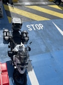 A motorcycle equipped with multiple storage boxes is parked on a brightly colored surface with the word 'STOP' painted in white. The surface features a mix of blue, yellow, and black diagonal stripes. Part of a red object with a rope can be seen on the left.