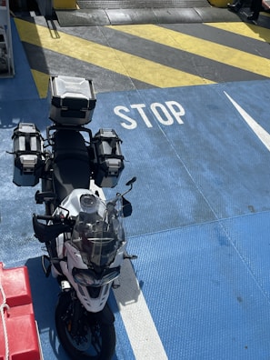A motorcycle equipped with multiple storage boxes is parked on a brightly colored surface with the word 'STOP' painted in white. The surface features a mix of blue, yellow, and black diagonal stripes. Part of a red object with a rope can be seen on the left.