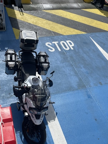 A motorcycle equipped with multiple storage boxes is parked on a brightly colored surface with the word 'STOP' painted in white. The surface features a mix of blue, yellow, and black diagonal stripes. Part of a red object with a rope can be seen on the left.