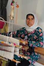 An older woman wearing a floral-patterned dress and a white headscarf sits in front of a traditional handloom. She is engaged in the process of weaving, surrounded by threads of various colors. Her expression is calm and focused.
