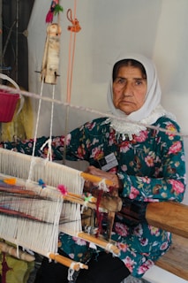 An older woman wearing a floral-patterned dress and a white headscarf sits in front of a traditional handloom. She is engaged in the process of weaving, surrounded by threads of various colors. Her expression is calm and focused.