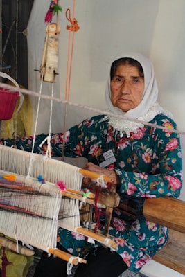 An older woman wearing a floral-patterned dress and a white headscarf sits in front of a traditional handloom. She is engaged in the process of weaving, surrounded by threads of various colors. Her expression is calm and focused.