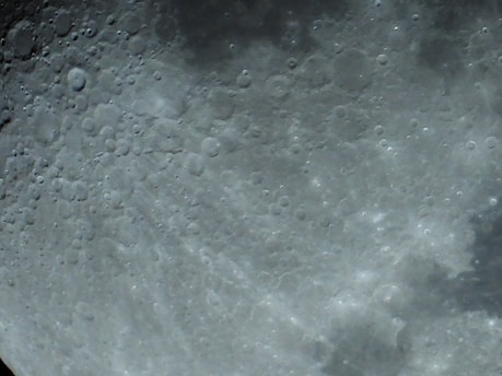 A detailed close-up of the moon's surface showing numerous craters and a textured, rocky landscape. The surface appears uneven and pockmarked, highlighting the rugged terrain.
