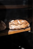 A loaf of rustic artisanal bread rests on a wooden shelf, partially illuminated by natural light. The crust is golden brown with a dusting of flour, creating a textured appearance. The surrounding area is in shadow, enhancing the focus on the bread.