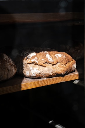 A loaf of rustic artisanal bread rests on a wooden shelf, partially illuminated by natural light. The crust is golden brown with a dusting of flour, creating a textured appearance. The surrounding area is in shadow, enhancing the focus on the bread.