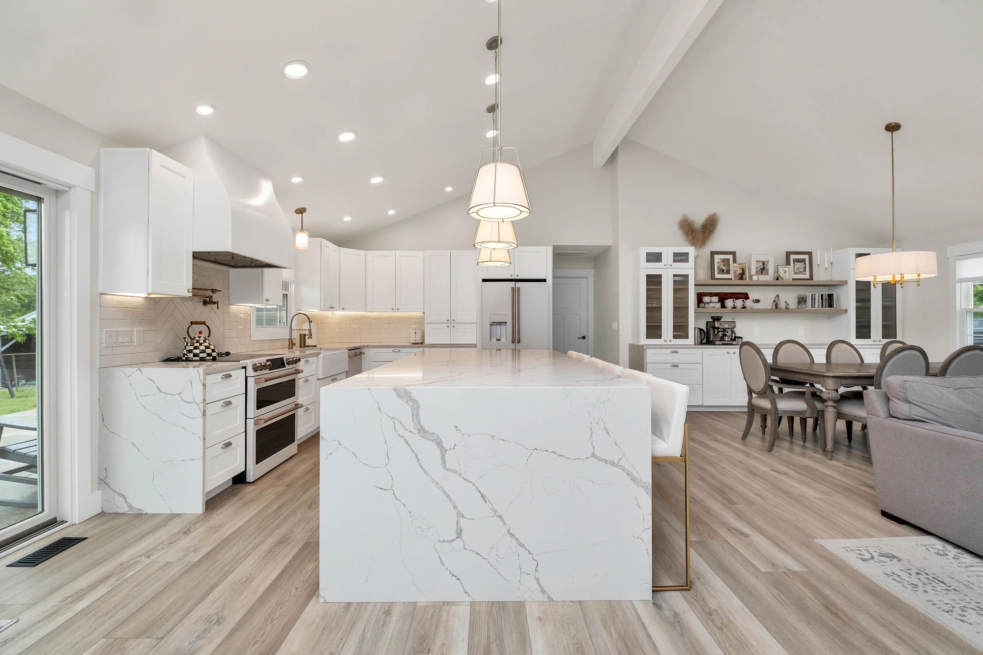 A bright, completed open-plan kitchen with white cabinetry, a large marble-look island, pendant lights, timber flooring, and an adjoining dining area.