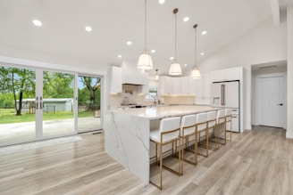 A modern kitchen with white cabinetry and a large marble island featuring seating for six. Pendant lights hang above the island, providing a warm ambiance. The kitchen is equipped with stainless steel appliances, including a refrigerator. Large windows and sliding doors offer a view of a lush green backyard, allowing natural light to brighten the space.