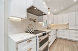 A modern kitchen with white cabinetry and brass hardware. The backsplash features herringbone design tiles, and a checkered kettle is placed on a sleek stovetop. The countertops are marble, and under-cabinet lighting illuminates the workspace. Light wood flooring adds warmth to the space.