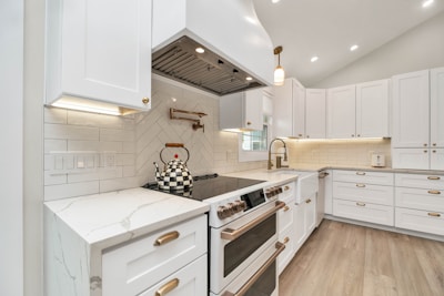 A modern kitchen with white cabinetry and brass hardware. The backsplash features herringbone design tiles, and a checkered kettle is placed on a sleek stovetop. The countertops are marble, and under-cabinet lighting illuminates the workspace. Light wood flooring adds warmth to the space.
