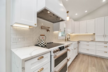 A modern kitchen with white cabinetry and brass hardware. The backsplash features herringbone design tiles, and a checkered kettle is placed on a sleek stovetop. The countertops are marble, and under-cabinet lighting illuminates the workspace. Light wood flooring adds warmth to the space.