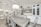 A modern kitchen and dining area with white cabinetry and light wood flooring. The dining table, set with six elegant chairs, is complemented by a large, decorative chandelier above. The kitchen features a white marble backsplash, a built-in stove, and an island with gold-accented bar stools. Shelving on the wall above the cabinets holds framed photos and decorative items, creating a cozy atmosphere.