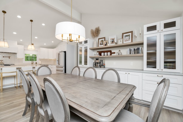 A modern kitchen and dining area with white cabinetry and light wood flooring. The dining table, set with six elegant chairs, is complemented by a large, decorative chandelier above. The kitchen features a white marble backsplash, a built-in stove, and an island with gold-accented bar stools. Shelving on the wall above the cabinets holds framed photos and decorative items, creating a cozy atmosphere.