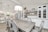 A modern kitchen and dining area with white cabinetry and light wood flooring. The dining table, set with six elegant chairs, is complemented by a large, decorative chandelier above. The kitchen features a white marble backsplash, a built-in stove, and an island with gold-accented bar stools. Shelving on the wall above the cabinets holds framed photos and decorative items, creating a cozy atmosphere.