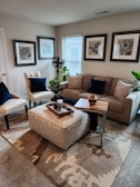 A cozy, well-decorated living room featuring a beige sectional sofa with patterned throw pillows and dark blue pillows, two white armchairs with blue accent pillows, and an upholstered ottoman with script writing. A wooden tray with decor items sits on the ottoman, and a small wooden side table is placed next to the sofa. Framed botanical artwork hangs on the walls above the sofa. There is a small potted plant on a stand next to the sofa and a larger plant by the window. A patterned area rug covers the floor.