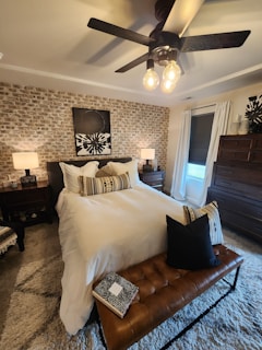 A cozy bedroom featuring a white bed with decorative cushions and a leather bench at the foot. The wall behind the bed is accented with a textured brick pattern and an abstract artwork. Warm lighting comes from modern ceiling lights and two bedside lamps. There is a large window with white curtains, and a wooden dresser stands in the corner.