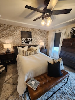 A cozy bedroom featuring a white bed with decorative cushions and a leather bench at the foot. The wall behind the bed is accented with a textured brick pattern and an abstract artwork. Warm lighting comes from modern ceiling lights and two bedside lamps. There is a large window with white curtains, and a wooden dresser stands in the corner.