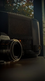 A vintage radio sits alongside an analog camera, both resting on a wooden surface. The background features blurry autumn leaves visible through a window, creating an ambiance of nostalgia.