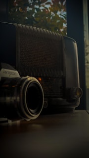 A vintage radio sits alongside an analog camera, both resting on a wooden surface. The background features blurry autumn leaves visible through a window, creating an ambiance of nostalgia.