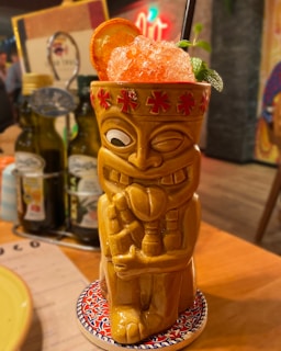 A tiki-style ceramic mug shaped like a character is filled with a crushed ice cocktail. The mug features red flower motifs around the rim. An orange slice and mint garnish the top of the drink with a straw inserted. The mug is placed on a colorful, patterned coaster on a wooden table, with olive oil bottles and menu items blurred in the background.