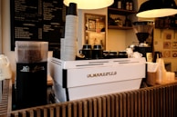A coffee shop interior featuring a modern espresso machine with a stack of paper cups on top. To the left, there is a grinder filled with coffee beans and a milk frother. In the background, a menu board displays various drink options, and nearby shelves hold jars and bottles. Warm lighting hangs overhead, adding to the cozy ambiance.