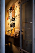 A narrow entryway filled with neatly arranged magazines and brochures on a wooden shelf. The wall features several framed posters, mostly promoting travel and lifestyle content. The ambient lighting casts a warm glow, creating a cozy atmosphere.