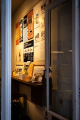 A narrow entryway filled with neatly arranged magazines and brochures on a wooden shelf. The wall features several framed posters, mostly promoting travel and lifestyle content. The ambient lighting casts a warm glow, creating a cozy atmosphere.