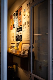 A narrow entryway filled with neatly arranged magazines and brochures on a wooden shelf. The wall features several framed posters, mostly promoting travel and lifestyle content. The ambient lighting casts a warm glow, creating a cozy atmosphere.