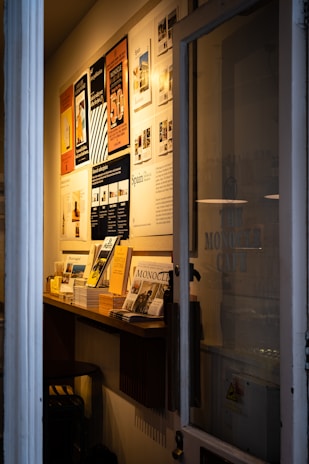 A narrow entryway filled with neatly arranged magazines and brochures on a wooden shelf. The wall features several framed posters, mostly promoting travel and lifestyle content. The ambient lighting casts a warm glow, creating a cozy atmosphere.