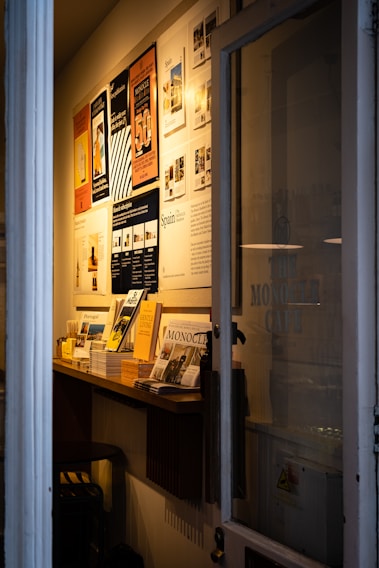 A narrow entryway filled with neatly arranged magazines and brochures on a wooden shelf. The wall features several framed posters, mostly promoting travel and lifestyle content. The ambient lighting casts a warm glow, creating a cozy atmosphere.