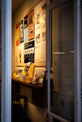 A narrow entryway filled with neatly arranged magazines and brochures on a wooden shelf. The wall features several framed posters, mostly promoting travel and lifestyle content. The ambient lighting casts a warm glow, creating a cozy atmosphere.