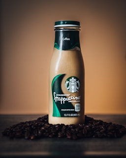 A bottle of Starbucks Frappuccino chilled coffee drink is placed on a surface surrounded by coffee beans. The background is a gradient of warm brown tones, creating a cozy and inviting atmosphere. The bottle is tall and slender with a green and white Starbucks logo prominently displayed.