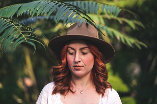 A woman with red hair is wearing a brown wide-brimmed hat. She is surrounded by green foliage, suggesting a natural or outdoor setting. Her expression is calm and serene, and she is wearing a white top with a small pendant necklace.