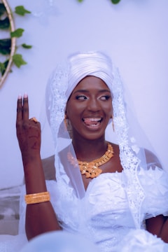 A person dressed in traditional wedding attire with a white headpiece and veil, adorned with gold jewelry. Their henna-decorated hand is raised with fingers forming a sign, and they have a joyful expression, sticking out their tongue slightly.