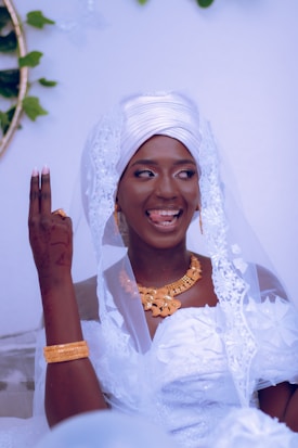 A person dressed in traditional wedding attire with a white headpiece and veil, adorned with gold jewelry. Their henna-decorated hand is raised with fingers forming a sign, and they have a joyful expression, sticking out their tongue slightly.