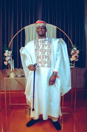A person dressed in traditional attire featuring a white flowing robe with intricate patterns, a matching cap, and holding a cane. The background includes floral arrangements and a decorative gold hoop, with dark curtains creating a formal setting.
