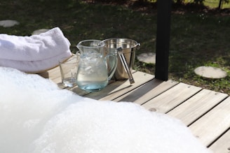 A wooden deck features a few neatly stacked white towels, a glass pitcher filled with ice water, a glass, and a metal ice bucket with tongs. The deck overlooks a grassy area with stepping stones, creating a serene and relaxing outdoor setting.