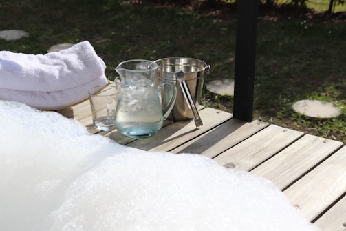 A wooden deck features a few neatly stacked white towels, a glass pitcher filled with ice water, a glass, and a metal ice bucket with tongs. The deck overlooks a grassy area with stepping stones, creating a serene and relaxing outdoor setting.