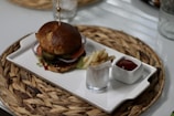 A gourmet hamburger with layers of sliced onions, pickles, and tomatoes sits on a rectangular white plate. Next to the burger is a small glass containing fries, accompanied by a square dish of ketchup. The plate is placed on a woven straw mat on a table.
