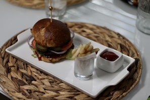 A gourmet hamburger with layers of sliced onions, pickles, and tomatoes sits on a rectangular white plate. Next to the burger is a small glass containing fries, accompanied by a square dish of ketchup. The plate is placed on a woven straw mat on a table.