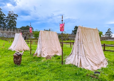 Three beige canvas tents stand on lush green grass with a stone fortress wall and trees in the background. A few medieval-style banners, spear-like poles, and a wood pile suggest a historical or reenactment scene.