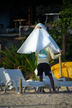 A man is standing on a sandy beach, holding a large white cloth or towel over his head. He is wearing a light-colored long-sleeve shirt and dark shorts, and appears to be near some lounge chairs arranged in pairs. The background shows green tropical plants and some wooden structures, with a yellow kayak partially visible.
