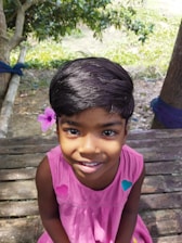 A young child with short black hair and a pink flower tucked behind the ear. The child is wearing a sleeveless pink dress adorned with colorful patches. The background features wooden planks and some green trees in a natural outdoor setting with sunlight casting shadows.