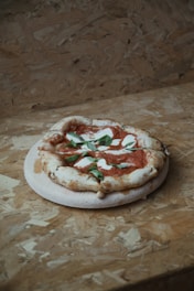 A rustic-style pizza with a slightly irregular shape sits on a wooden board. The pizza is topped with tomato sauce, fresh basil leaves, and mozzarella cheese. The background consists of a textured, unfinished plywood surface.