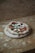 A rustic-style pizza with a slightly irregular shape sits on a wooden board. The pizza is topped with tomato sauce, fresh basil leaves, and mozzarella cheese. The background consists of a textured, unfinished plywood surface.