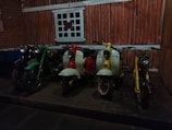 A row of vintage scooters and motorcycles is positioned in front of a rustic, wood-paneled wall. The scene includes a green motorcycle, a red and white scooter, a second white scooter with yellow accents, and a yellow scooter. The floor is stone, and there is a window with a grid design above the vehicles.