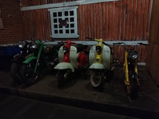 A row of vintage scooters and motorcycles is positioned in front of a rustic, wood-paneled wall. The scene includes a green motorcycle, a red and white scooter, a second white scooter with yellow accents, and a yellow scooter. The floor is stone, and there is a window with a grid design above the vehicles.