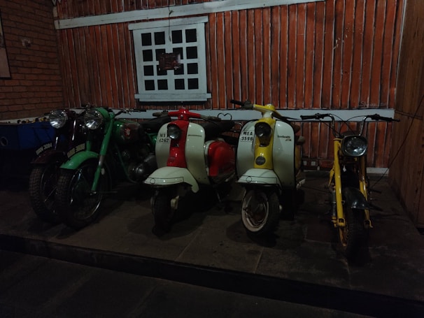 A row of vintage scooters and motorcycles is positioned in front of a rustic, wood-paneled wall. The scene includes a green motorcycle, a red and white scooter, a second white scooter with yellow accents, and a yellow scooter. The floor is stone, and there is a window with a grid design above the vehicles.