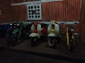 A row of vintage scooters and motorcycles is positioned in front of a rustic, wood-paneled wall. The scene includes a green motorcycle, a red and white scooter, a second white scooter with yellow accents, and a yellow scooter. The floor is stone, and there is a window with a grid design above the vehicles.