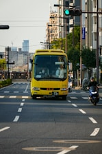 A bright yellow bus is driving down a city street with trees and buildings on either side. It is accompanied by a motorcyclist wearing a helmet on the adjacent lane. The road is marked with white dashed lines, and a green traffic light is visible in the background. The scene is set during the day with clear skies.